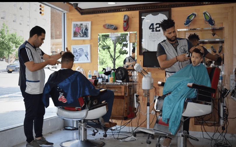 Two barbers cutting hair for a man and a yawning child in a sports-themed barbershop.