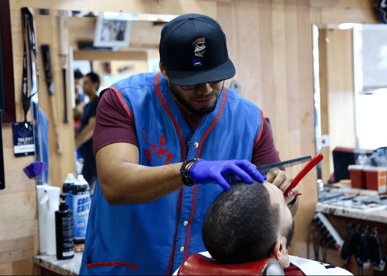 Barber in a blue vest trims a man's hair with a straight razor.